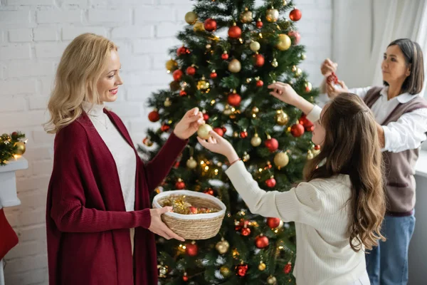 Adolescent fille donnant boule à sourire mère tout en décorant arbre de Noël près de mamie multiraciale — Photo de stock