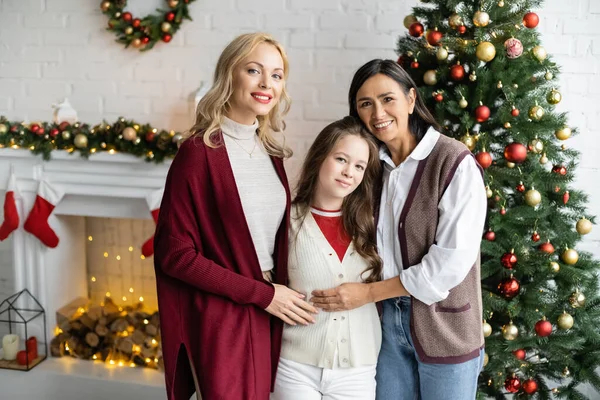 Mujer rubia feliz mirando a la cámara cerca de la hija y la mamá multirracial en la sala de estar con decoración de Navidad - foto de stock