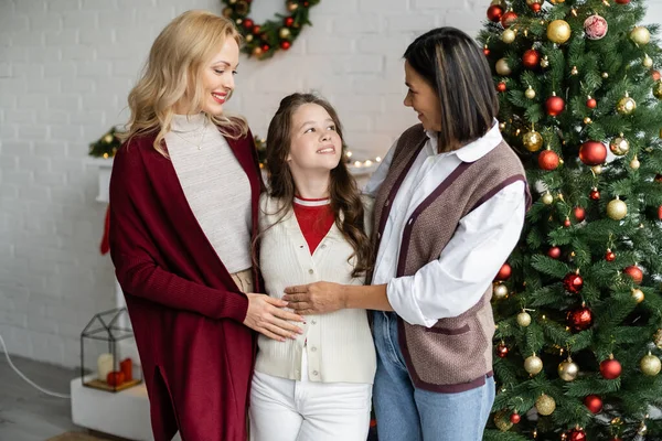 Веселая девочка-подросток улыбается рядом с матерью и многорасовой бабушкой возле рождественской елки — стоковое фото