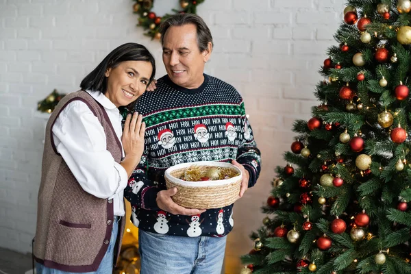 Glücklich multirassische Frau blickt in die Kamera in der Nähe Ehemann mit Weihnachtskugeln in Weidenkorb — Stockfoto