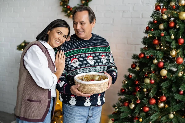 Happy multiracial woman looking at camera near husband with christmas balls in wicker basket — Stock Photo