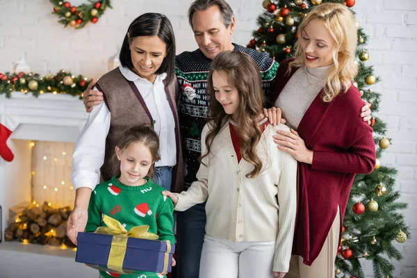 Preteen Mädchen hält Geschenkbox in der Nähe lächelnde multiethnische Familie im Wohnzimmer mit Weihnachtsdekoration — Stockfoto
