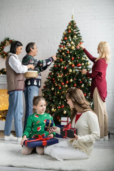 Schwestern sitzen auf dem Boden mit Neujahrsgeschenken, während multiethnische Familie den Weihnachtsbaum schmückt — Stockfoto