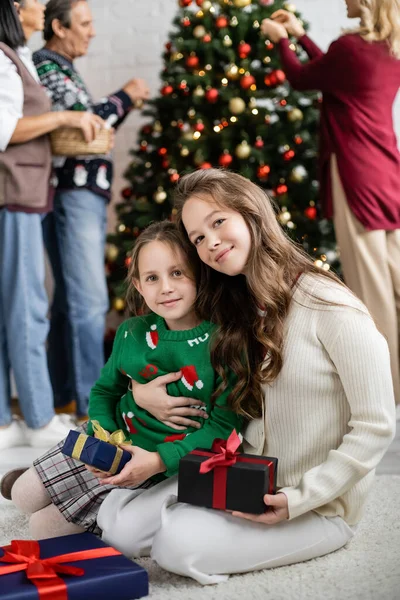 Preteen fille étreinte soeur près de boîtes-cadeaux et multiethnique famille décoration arbre de Noël sur fond flou — Photo de stock