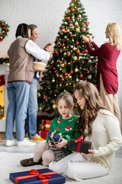 Blurred multicultural family decorating christmas tree near girls sitting on floor with gift boxes — Stock Photo