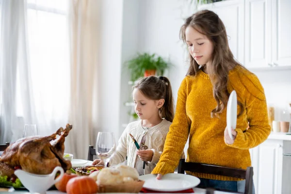 Hermanas sosteniendo platos y cubiertos mientras sirven mesa para la cena de Acción de Gracias cerca de pavo asado - foto de stock