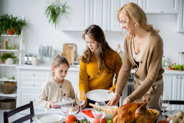 Mujer rubia sonriente sirviendo mesa para la cena de acción de gracias con hijas en la cocina - foto de stock