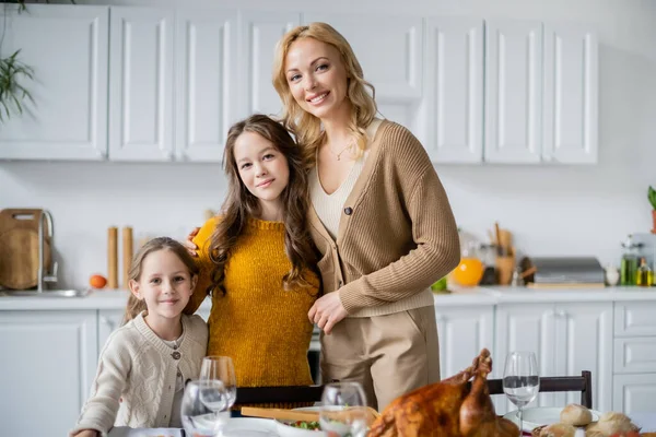 Blonde woman with daughters smiling at camera near thanksgiving dinner served in kitchen — Stock Photo
