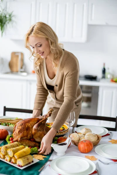 Feliz mujer rubia sirviendo mesa con pavo asado cerca de maíz a la parrilla y bollos para la cena festiva de acción de gracias - foto de stock