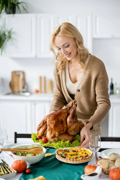 Sorrindo mulher loira segurando peru assado perto de torta de abóbora e salada de legumes servidos para o jantar de ação de graças — Fotografia de Stock