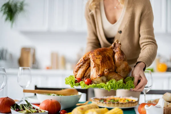 Vista parcial de la mujer sosteniendo delicioso pavo asado con verduras frescas cerca de la mesa con cena de acción de gracias - foto de stock