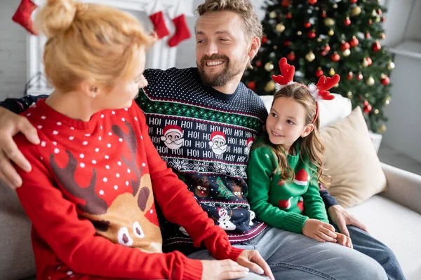 Улыбающийся мужчина обнимает жену и дочь в рождественских свитерах дома — стоковое фото