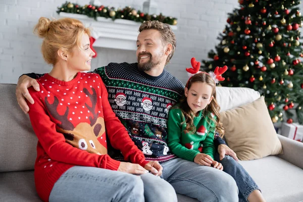 Fröhlicher Mann umarmt Tochter und Ehefrau im Weihnachtspullover auf der heimischen Couch — Stockfoto