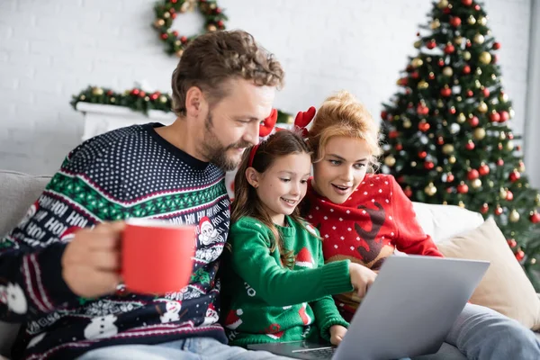 Chica sonriente usando el ordenador portátil con los padres en suéteres de Navidad en casa - foto de stock