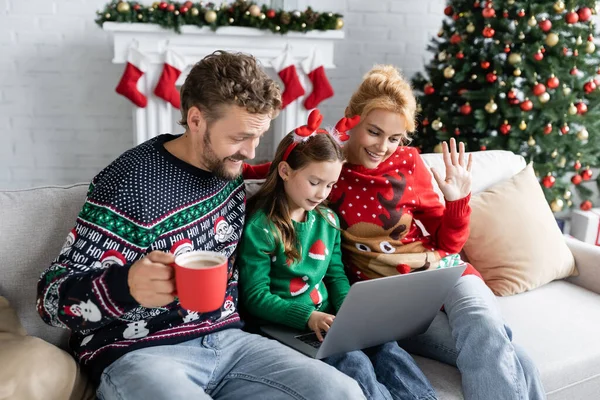 Donna sorridente in maglione di Natale che ha videochiamata sul computer portatile vicino alla famiglia sul divano a casa — Foto stock