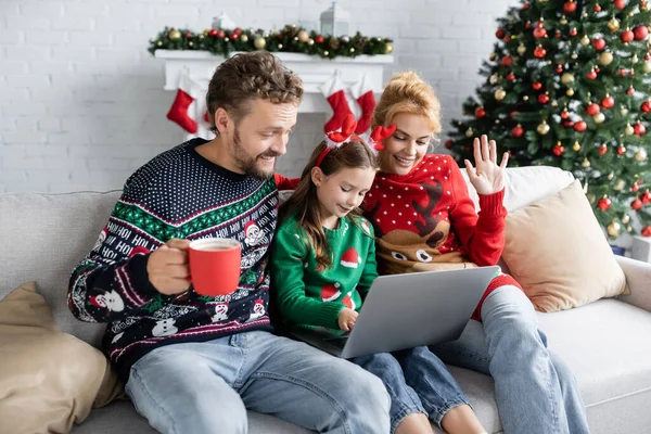 Famille souriante en chandails de nouvelle année ayant appel vidéo sur ordinateur portable à la maison — Photo de stock