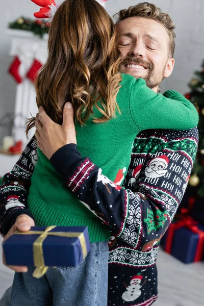 Hombre positivo en suéter abrazando hija y sosteniendo borrosa navidad presente en casa - foto de stock