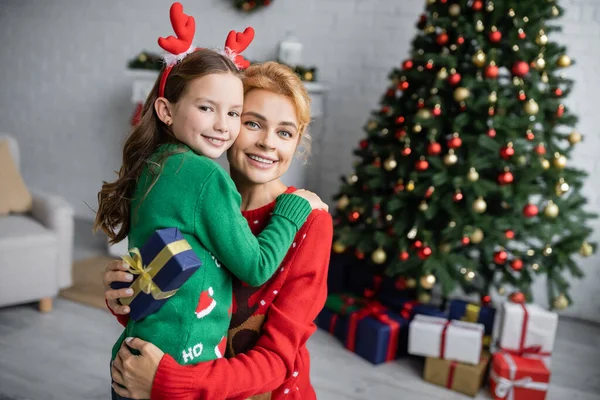 Mujer sonriente en suéter abrazando a su hija y sosteniendo el regalo de Navidad en casa - foto de stock