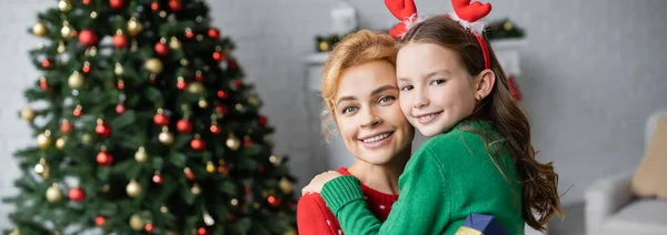 Femme heureuse en pull étreignant sa fille près de l'arbre de Noël flou à la maison, bannière — Photo de stock