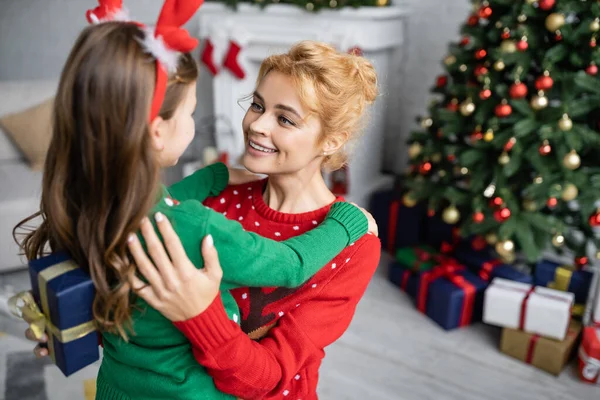 Glückliche Mutter umarmt Tochter im Pullover und hält Weihnachtsgeschenk zu Hause — Stockfoto