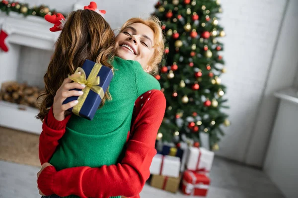 Sonriente mamá en suéter abrazando a la hija y la celebración de año nuevo presente en casa - foto de stock