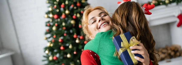 Lächelnde Eltern umarmen Tochter und halten Weihnachtsgeschenkbox zu Hause, Banner — Stockfoto