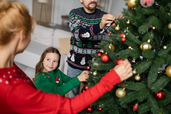 Criança sorridente em suéter decorando árvore de Natal com os pais em casa — Fotografia de Stock