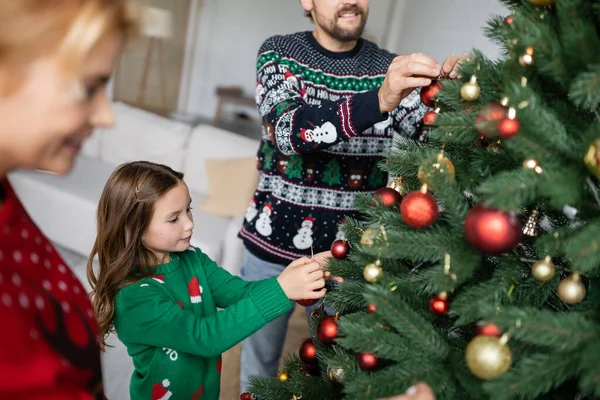 Menina decorando árvore de Natal com bolas perto de pais desfocados em casa — Fotografia de Stock