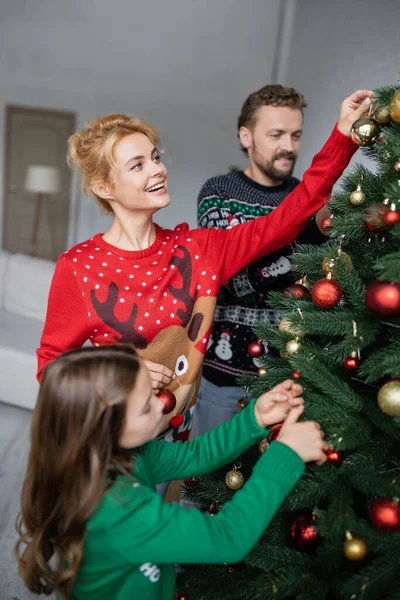 Счастливая женщина украшает елку шарами рядом с семьей в свитерах дома — стоковое фото