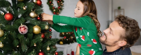 Father holding daughter in sweater decorating christmas tree at home, banner — Stock Photo