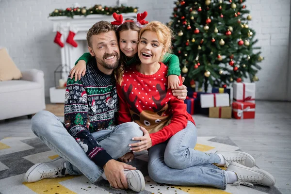 Mädchen umarmt Mutter und Vater in Weihnachtspullis und schaut zu Hause in die Kamera — Stock Photo