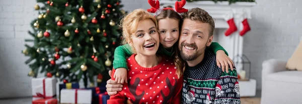 Веселая девушка обнимает отца и маму в теплых свитерах возле размытой рождественской елки дома, баннер — стоковое фото