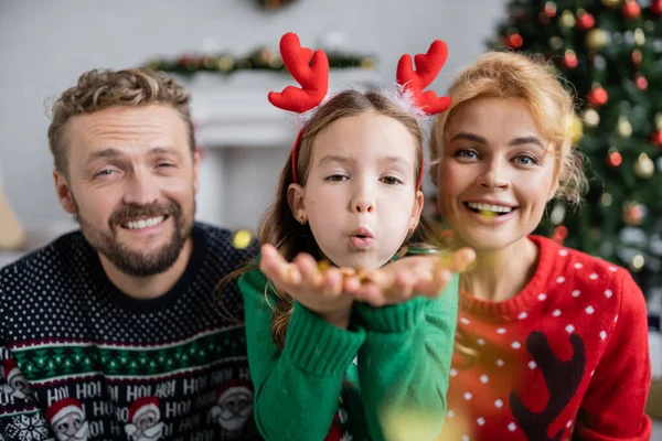 Chica soplando confeti en la cámara cerca de padres sonrientes en suéteres de Navidad en casa - foto de stock