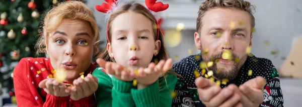 Criança e pais soprando confete na câmera durante a celebração do Natal em casa, banner — Fotografia de Stock