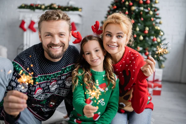 Cheerful family in christmas sweaters holding sparklers and looking at camera at home — Stock Photo