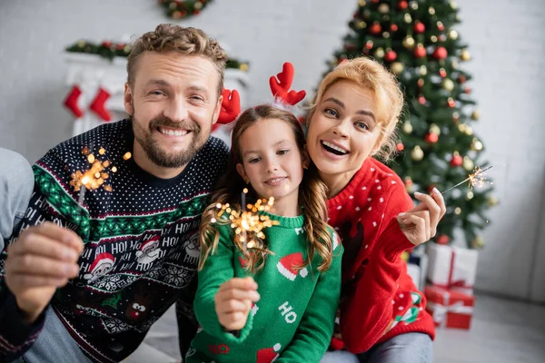 Famille positive dans des pulls chauds tenant des étincelles lors de la célébration de la nouvelle année à la maison — Photo de stock