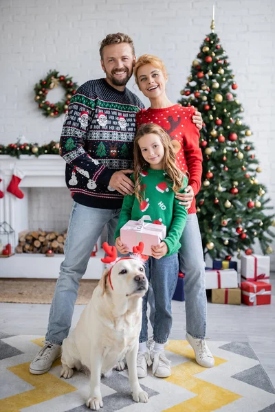 Smiling parents hugging near daughter with gift and labrador during christmas at home — Stock Photo