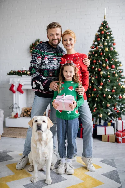 Famille heureuse avec cadeau regardant la caméra près de labrador pendant Noël à la maison — Photo de stock