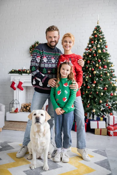 Famille multiethnique étreignant et regardant la caméra près du labrador pendant la nouvelle année à la maison — Photo de stock