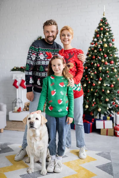 Sonrientes padres abrazando a hija cerca de labrador y borroso árbol de navidad en casa - foto de stock