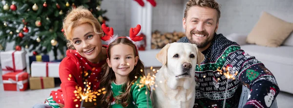 Familia feliz en jerseys navideños sosteniendo bengalas cerca de labrador en casa, pancarta - foto de stock