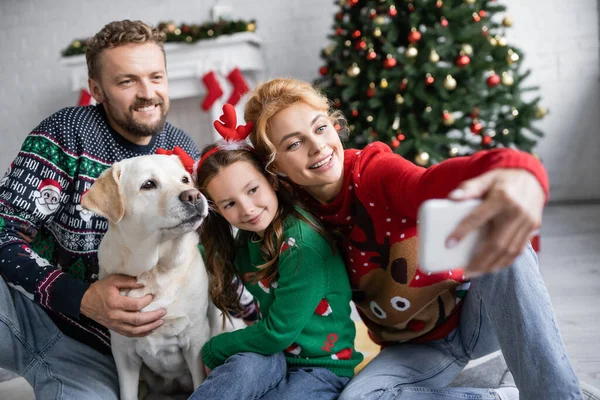 Mulher sorridente tomando selfie com a família e labrador durante o Natal em casa — Stock Photo