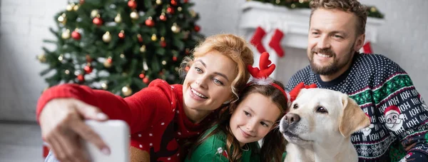 Família alegre com labrador tirando selfie no smartphone durante o ano novo em casa, banner — Fotografia de Stock