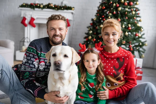 Fröhliche Familie in warmen Pullovern umarmt verschwommenen Labrador und blickt zu Hause in die Kamera — Stockfoto