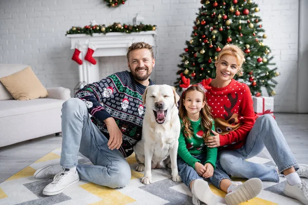 Família em blusas de Natal e jeans olhando para a câmera perto labrador em casa — Fotografia de Stock