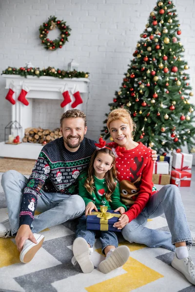 Familia positiva sosteniendo regalo de Navidad cerca de la decoración festiva en casa - foto de stock
