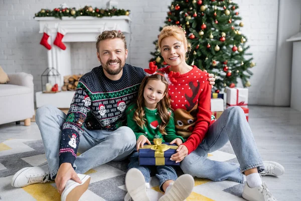 Famille joyeuse tenant cadeau et regardant la caméra pendant Noël à la maison — Photo de stock