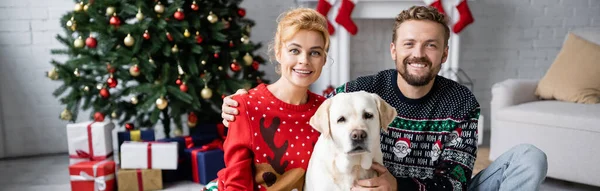 Casal sorridente em camisolas quentes de Natal olhando para a câmera perto labrador em casa, banner — Fotografia de Stock