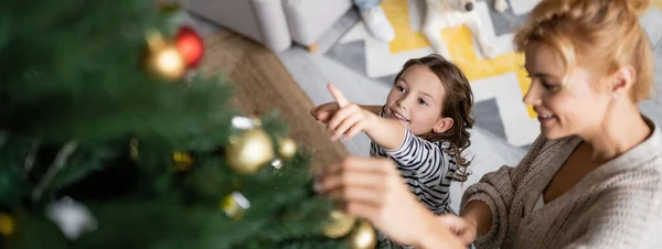 Vista de ángulo alto de la chica sonriente apuntando al árbol de Navidad cerca de mamá en casa, pancarta - foto de stock
