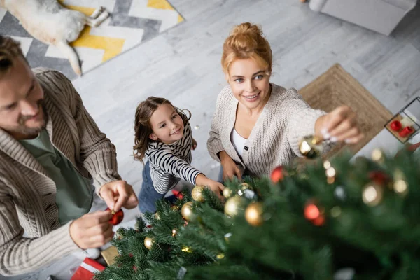 Вид под высоким углом улыбающейся семьи, держащей шарики у елки дома — стоковое фото